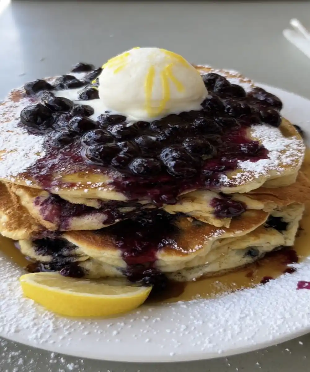 Stack of blueberry pancakes topped with lemon zest, whipped butter, and blueberry compote on white plate