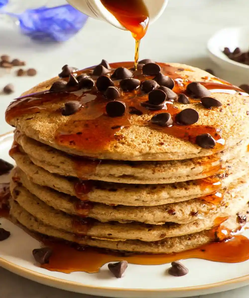 Stack of chocolate chip pancakes topped with syrup and chocolate chips being poured with maple syrup