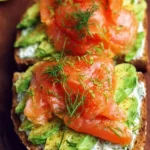 Avocado toast topped with smoked salmon and herbs on a rustic wooden table.